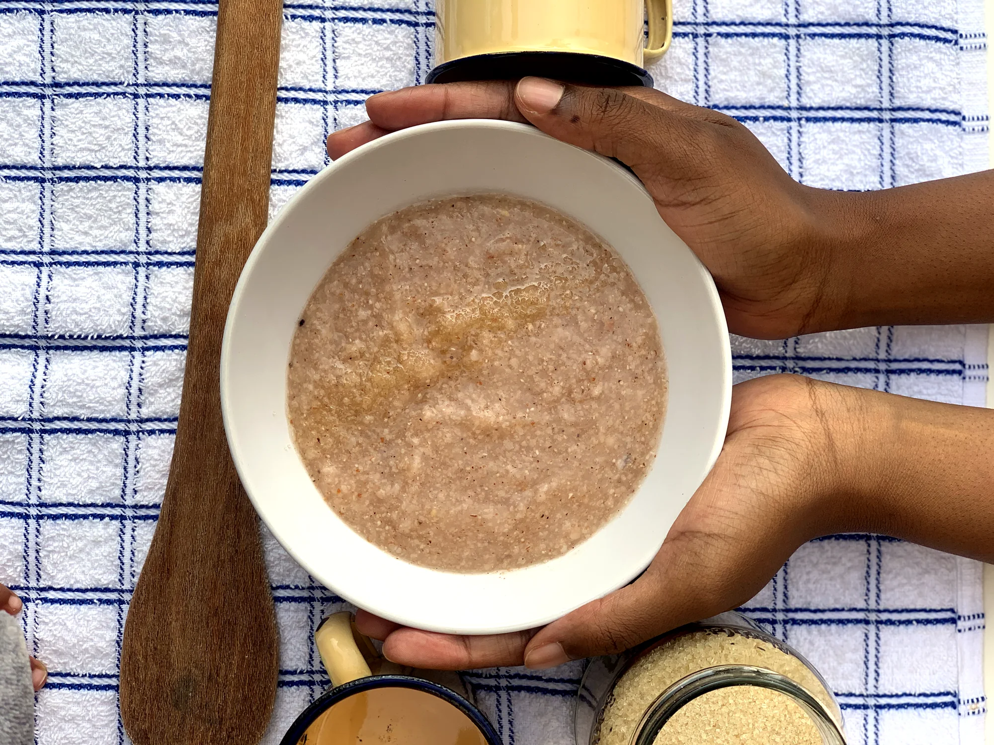 A bowl of Ting Ya Koko mabele held in two hands, with a wooden spoon and enamel cups on a navy-grid cloth